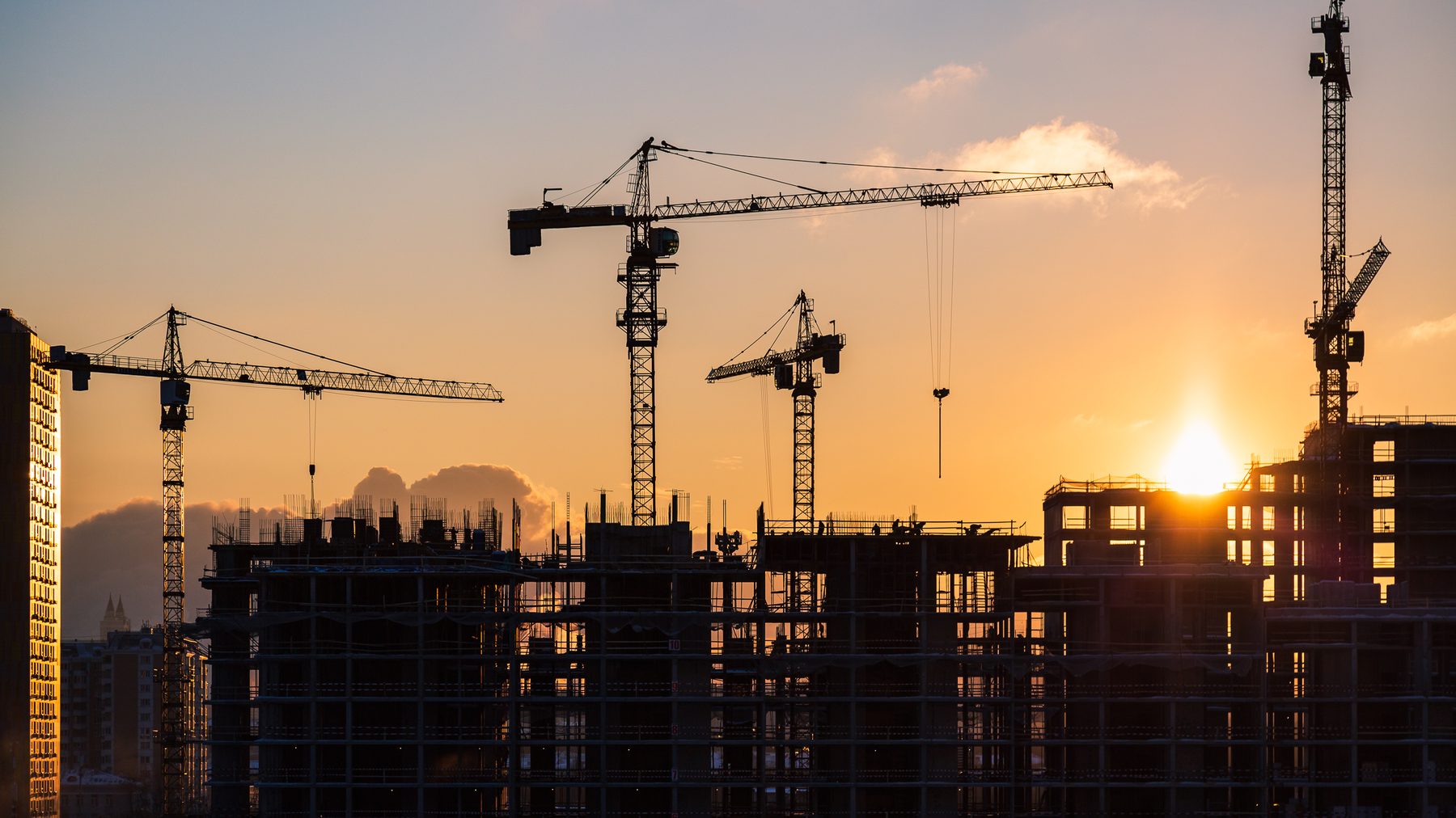 Tower cranes over buildings under construction, silhouetted against a sunset